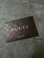 A dark brown Gucci leather card or plaque featuring the embossed GG monogram pattern and gold-toned 'GUCCI' lettering, placed on a textured gray surface.