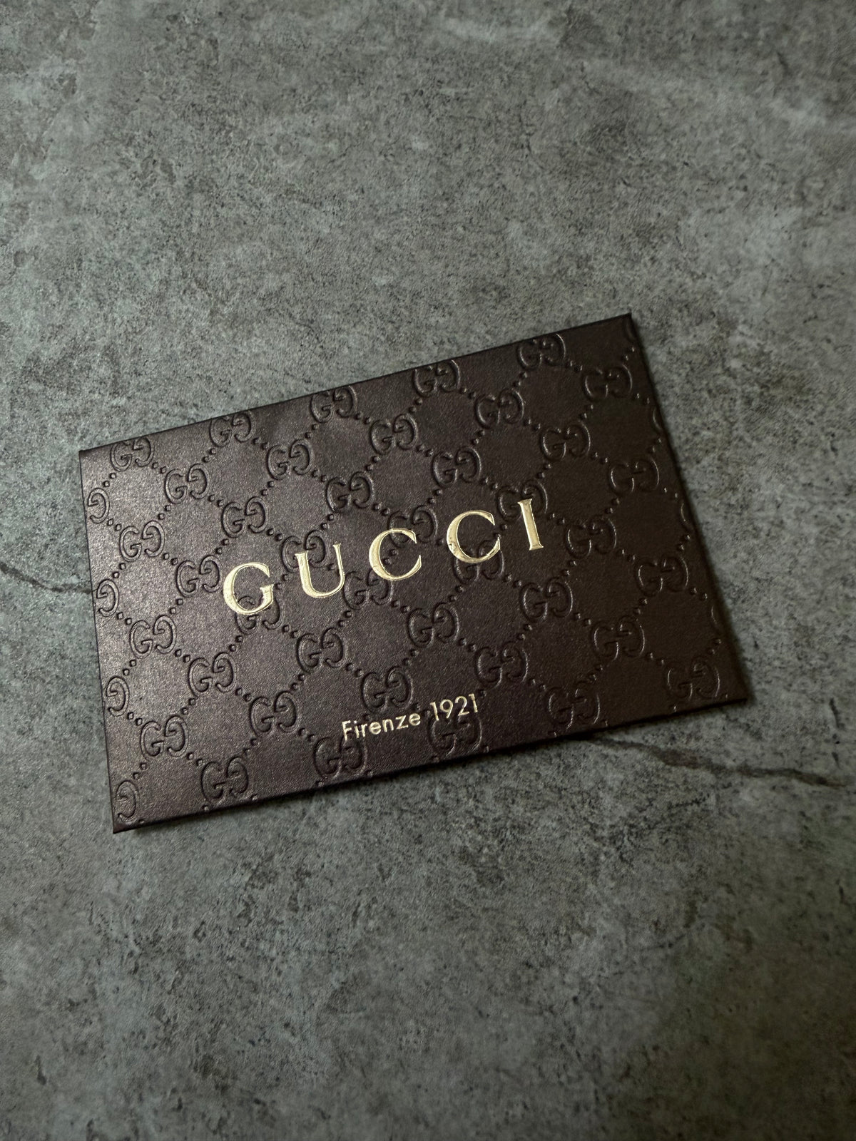 A dark brown Gucci leather card or plaque featuring the embossed GG monogram pattern and gold-toned 'GUCCI' lettering, placed on a textured gray surface.